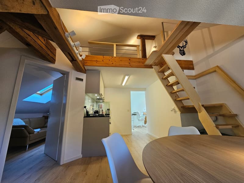 Open layout, wooden beam ceiling, modern staircase, white kitchen with countertops, living room with sofa and table, wooden floors, open door to loft area with couch and skylight