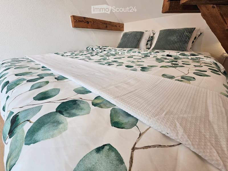 A cozy bedroom with a bed featuring a green leaf-patterned duvet and matching pillows, set against a white wall with wooden beams.