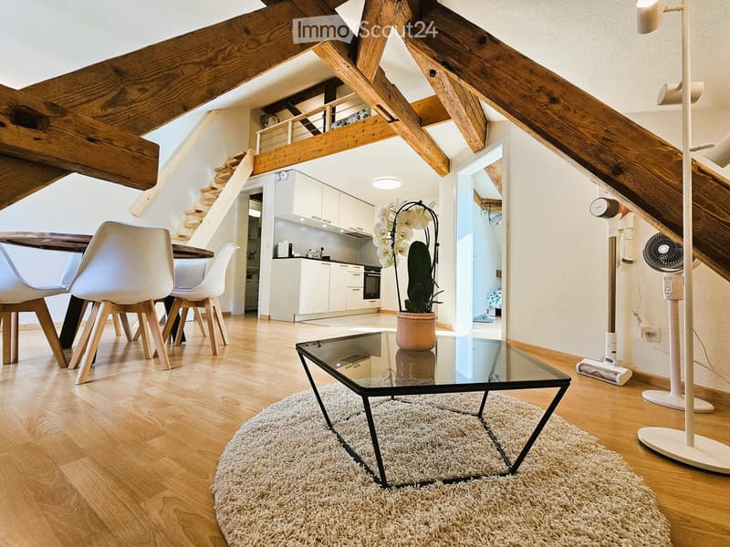 Modern living room with exposed wooden beams, white chairs, glass coffee table, and a small kitchen area visible in the background.