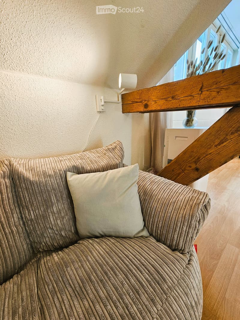 A cozy living room corner with a textured sofa, decorative pillows, a wall-mounted lamp, and a vase on a small table near a window with a wooden railing.