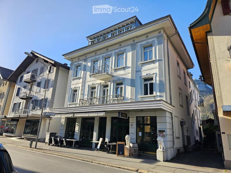 3-story building, white exterior, multiple windows, balconies, double front doors, lion statues at entrance, awnings over windows, table and chairs on sidewalk