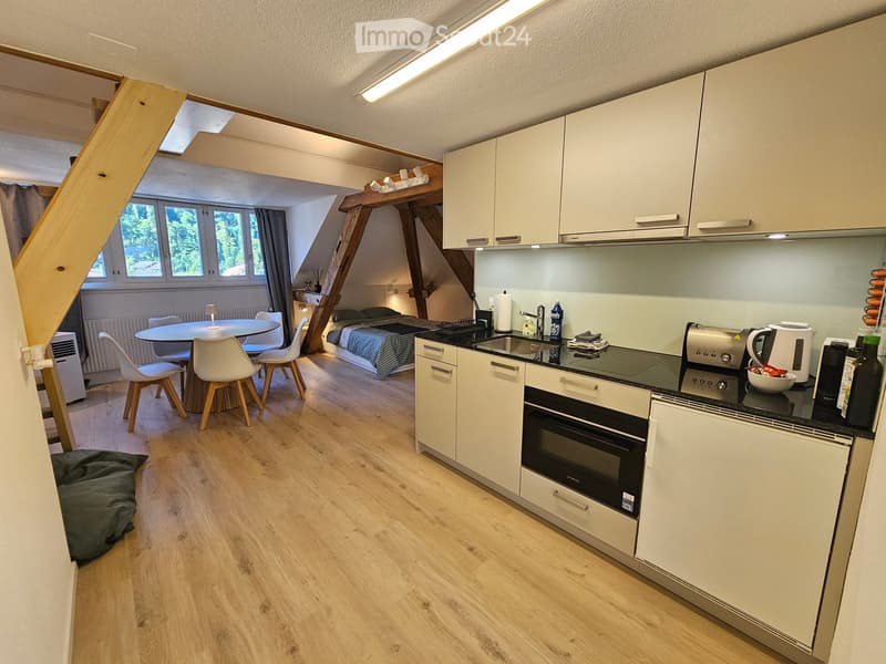 A kitchen with white cabinets, black countertops, wooden floor, table and chairs, an oven, a toaster, a kettle, and a bed under a wooden beam.