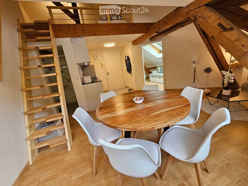 A modern, open-concept living and dining area with wooden floors, a round wooden dining table surrounded by white chairs, a small staircase leading to an upper loft area, and a small kitchen area visible in the background.