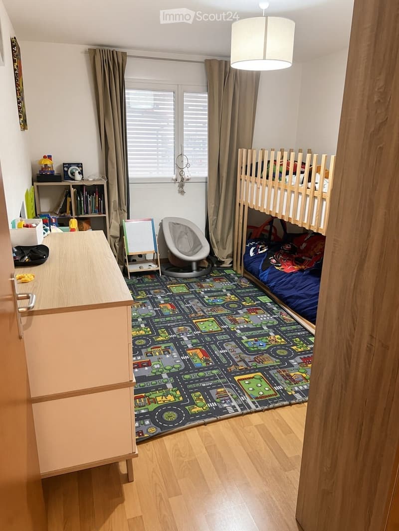 A cozy children's bedroom with a bunk bed, a colorful play mat, a small table, a chair, and a shelf with books and toys.