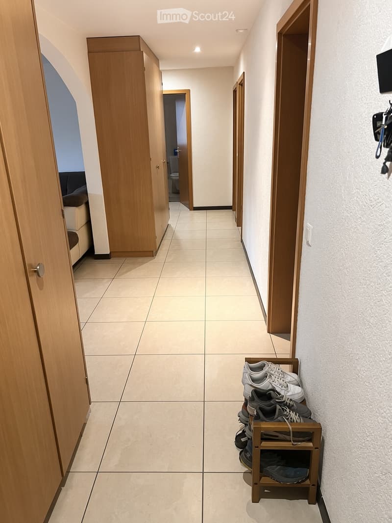 A clean, well-lit hallway with tiled flooring, wooden doors, and a small shoe rack holding several pairs of shoes. The hallway leads to other rooms, including a visible bathroom and a living area.