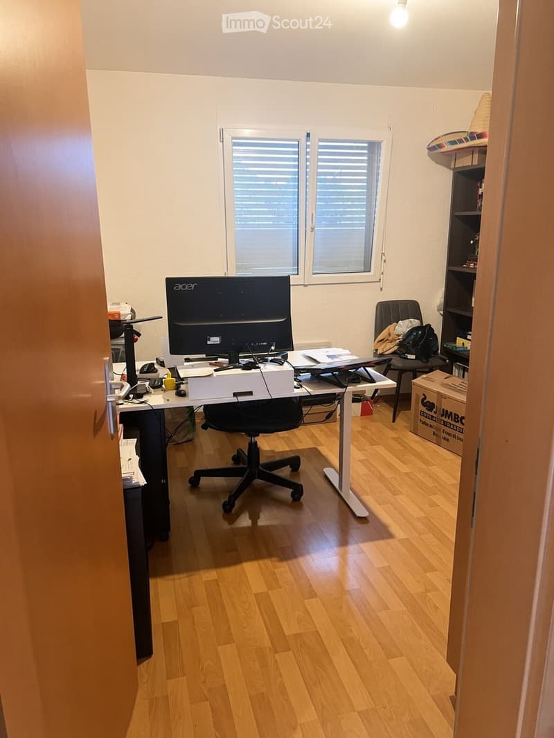 A furnished room with a wooden floor, a desk with a computer setup, a chair, and a shelf with various items.