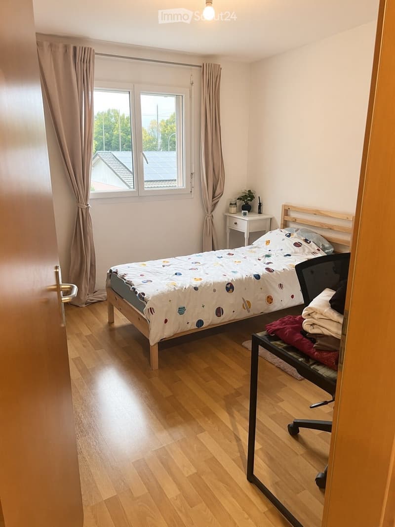 Cozy bedroom with wooden flooring, a bed with a colorful space-themed duvet, a nightstand with a plant, a chair, and a mirror.