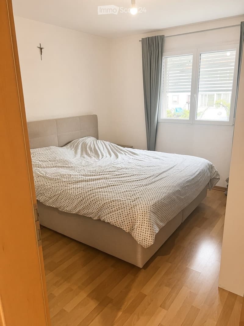 A modern bedroom with a large bed, wooden flooring, and large windows with grey curtains.