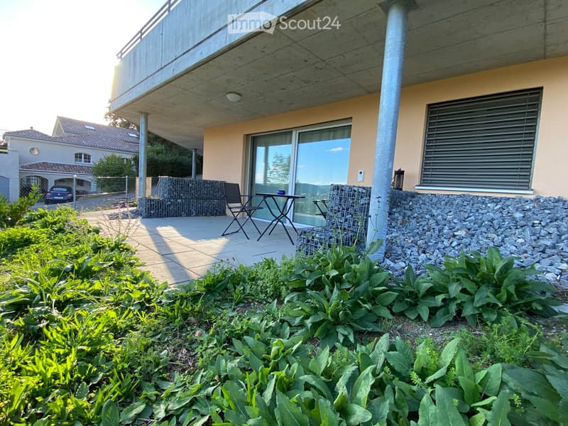 Garden area with lush greenery, a small patio with a table and chairs, and a modern building in the background.
