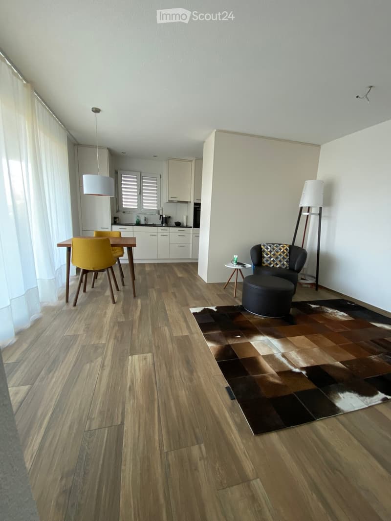 Open-plan living and dining area with wooden flooring, a patterned rug, a black leather sofa with an ottoman, a small side table, a dining table with yellow chairs, and a modern kitchen area with white cabinets and stainless steel appliances.