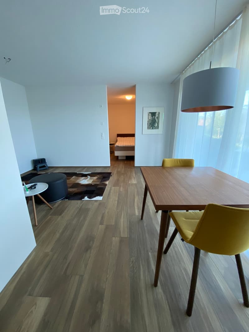 Modern open-plan living and dining area with wooden flooring, a dining table with chairs, a round stool, and a picture frame on the wall. A bedroom is visible in the background.