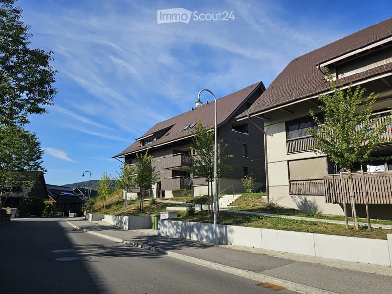 Two-story residential buildings with balconies, surrounded by trees and greenery, on a sunny day.