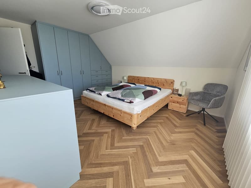 Cozy attic bedroom with wooden herringbone flooring, a large wooden bed with colorful bedding, a grey armchair, a nightstand with a lamp, and a tall grey wardrobe.