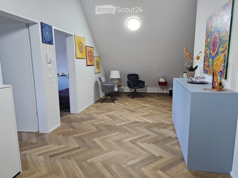 A modern, well-lit room with herringbone wooden flooring, two chairs, a small table, a lamp, and decorative artwork on the walls.