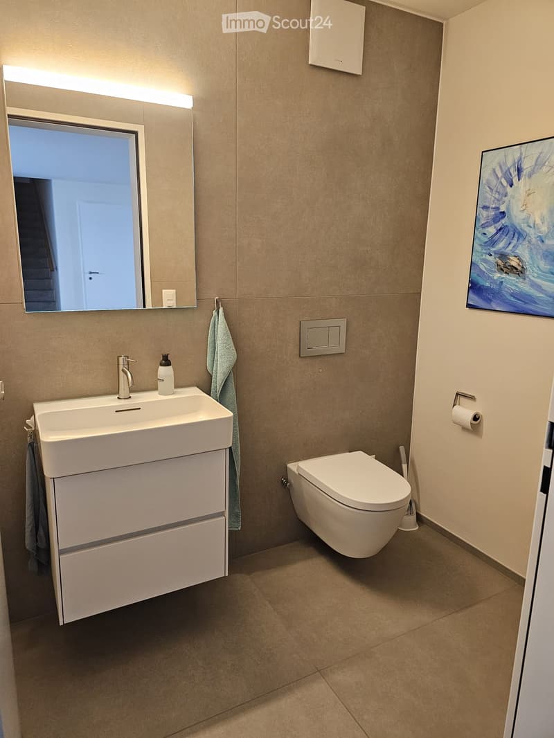 Modern bathroom with beige tiled walls, a white sink with a chrome faucet, a toilet, a large mirror with built-in lighting, and a towel hanging on a rack.