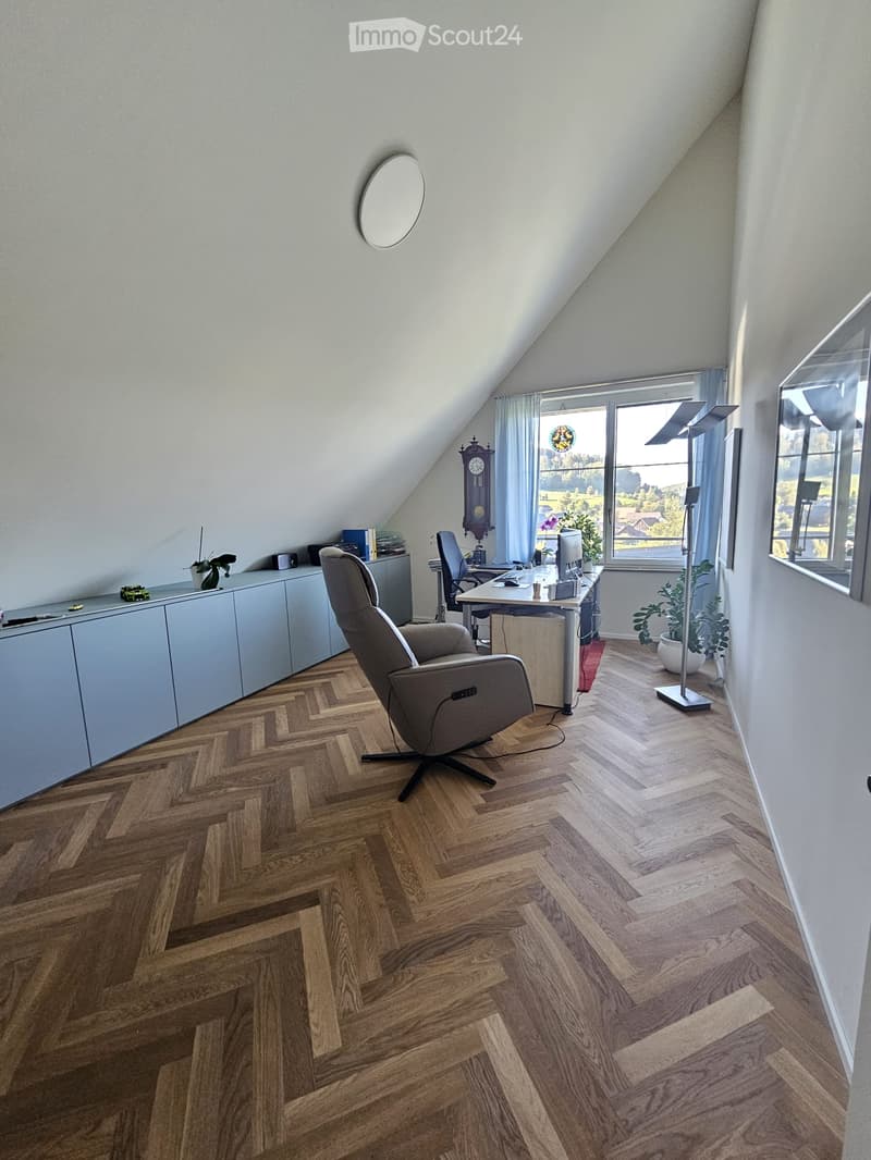 Bright attic office with herringbone wooden flooring, large window with view, grey office chair, desk with computer, cabinet, round mirror, floor lamp, and decorative items.