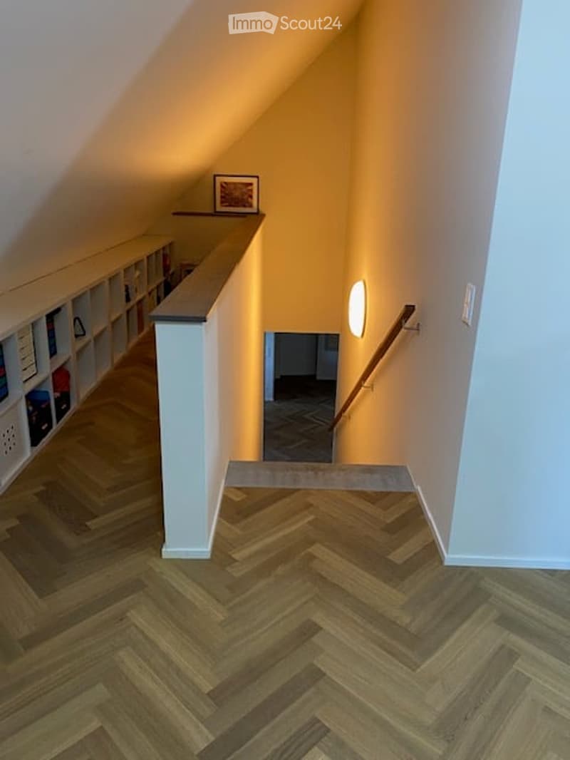 Spacious hallway with herringbone wooden flooring, white walls, built-in shelving on one side, and a small picture frame on the shelf. Warm lighting from a wall-mounted lamp.