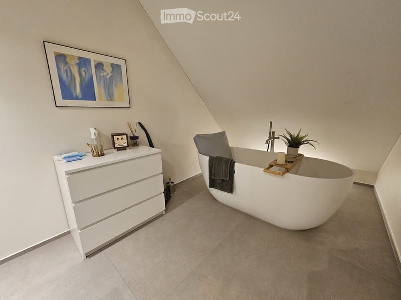 Modern bathroom with a freestanding bathtub, a white bathroom cabinet, a towel draped over the edge of the tub, a small plant, and a candle on a wooden tray. The walls are light-colored with a slanted ceiling typical of an attic space.