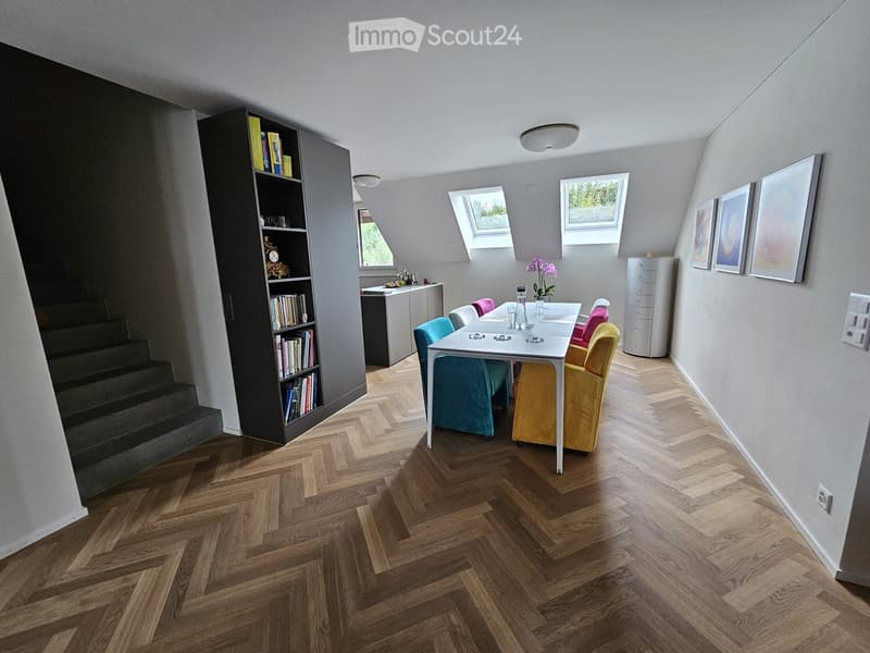 Bright, modern room with herringbone wooden flooring, white walls, and skylights. Features a dining table with chairs, a bookshelf, and a small kitchen area.