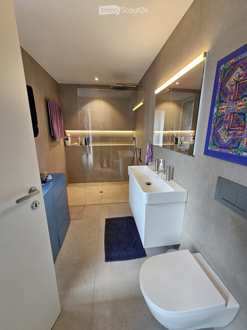 Modern bathroom with a white sink, bathtub, large mirror, and glass-enclosed shower area. Features include built-in lighting, a towel rack, and a decorative wall art piece.