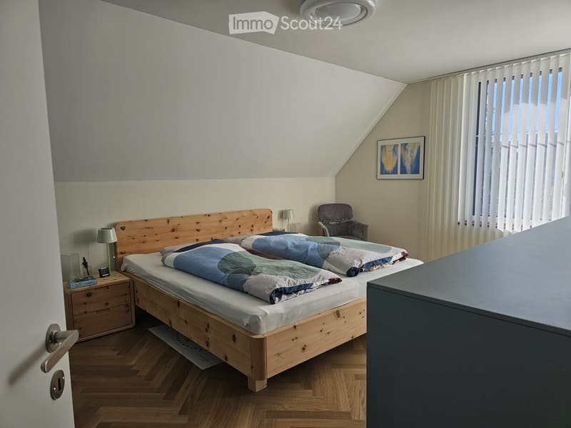 Cozy bedroom with a wooden bed, nightstand, chair, and artwork on the wall. The room features wooden flooring and large windows with blinds.