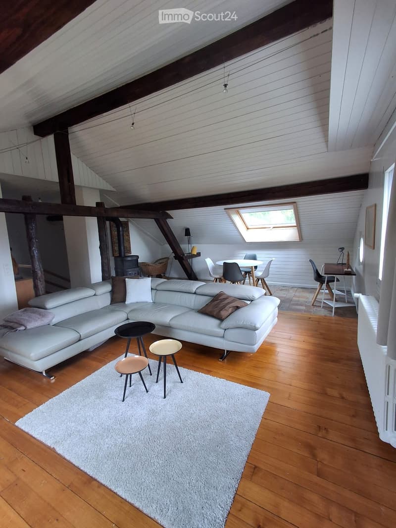 Bright, airy living space with exposed wooden beams, white shiplap walls, and wooden flooring. Features a white sectional sofa with throw pillows, a small black coffee table, and three small round stools on a white shag rug. A dining area with a round tab