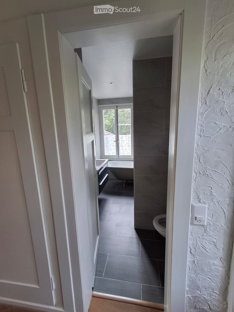 Modern bathroom with a bathtub, toilet, sink, and mirror. The room features grey tiles and a large window with a view of greenery outside.