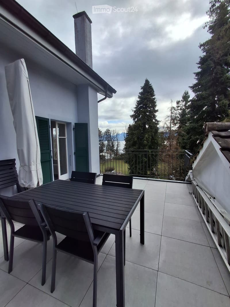 Balcony with a wooden table and four chairs, overlooking a scenic view of trees and a body of water.