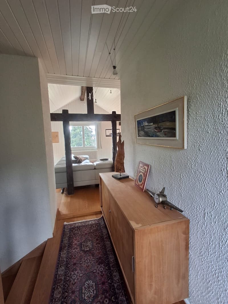 A narrow hallway with wooden flooring, a patterned rug, and a wooden side table with decorative items, leading to a living area with a sofa and artwork.