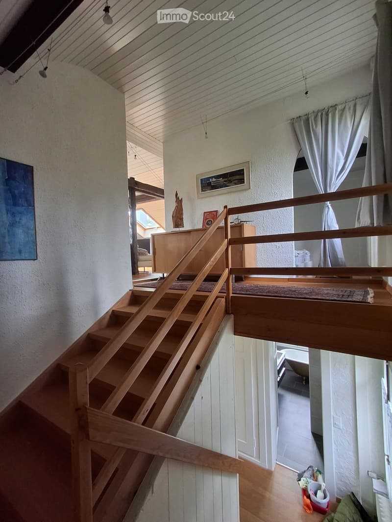 Wooden staircase leading to an upper level with a wooden railing, white walls, and a skylight above. The upper level has a small landing with a rug and a view of a room with a bed and a painting on the wall.