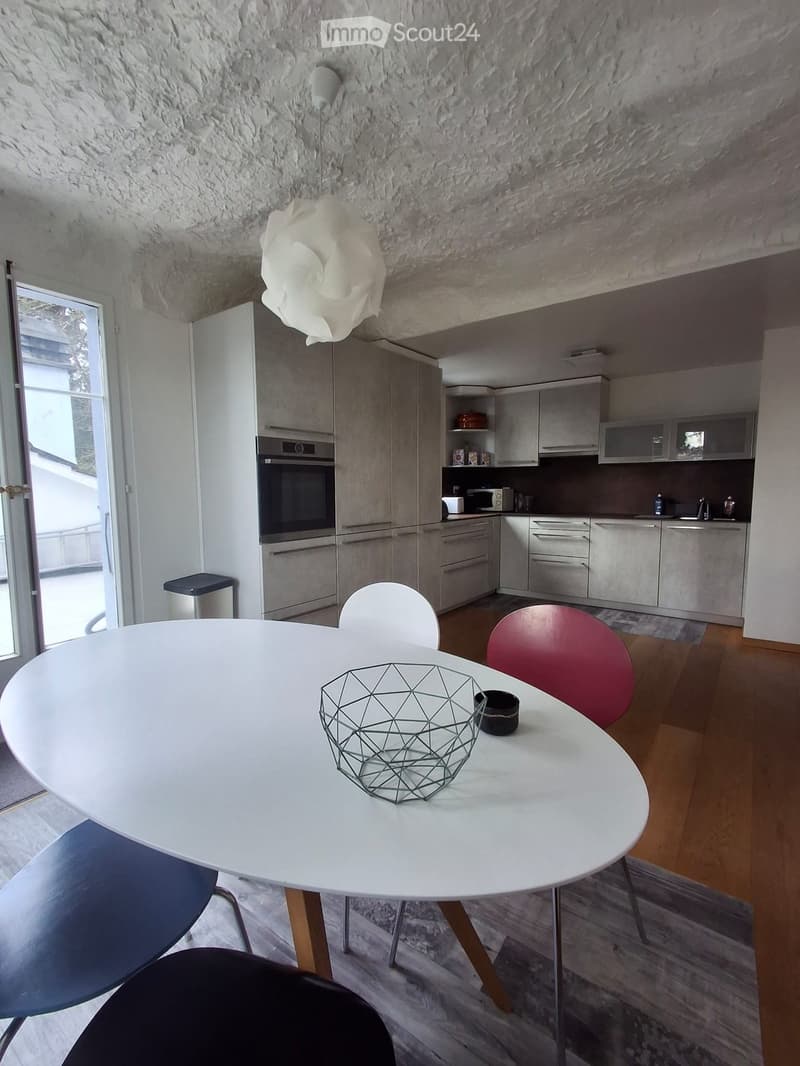 Modern kitchen and dining area with white cabinets, stainless steel appliances, a round white dining table with colorful chairs, and a geometric wire basket on the table.
