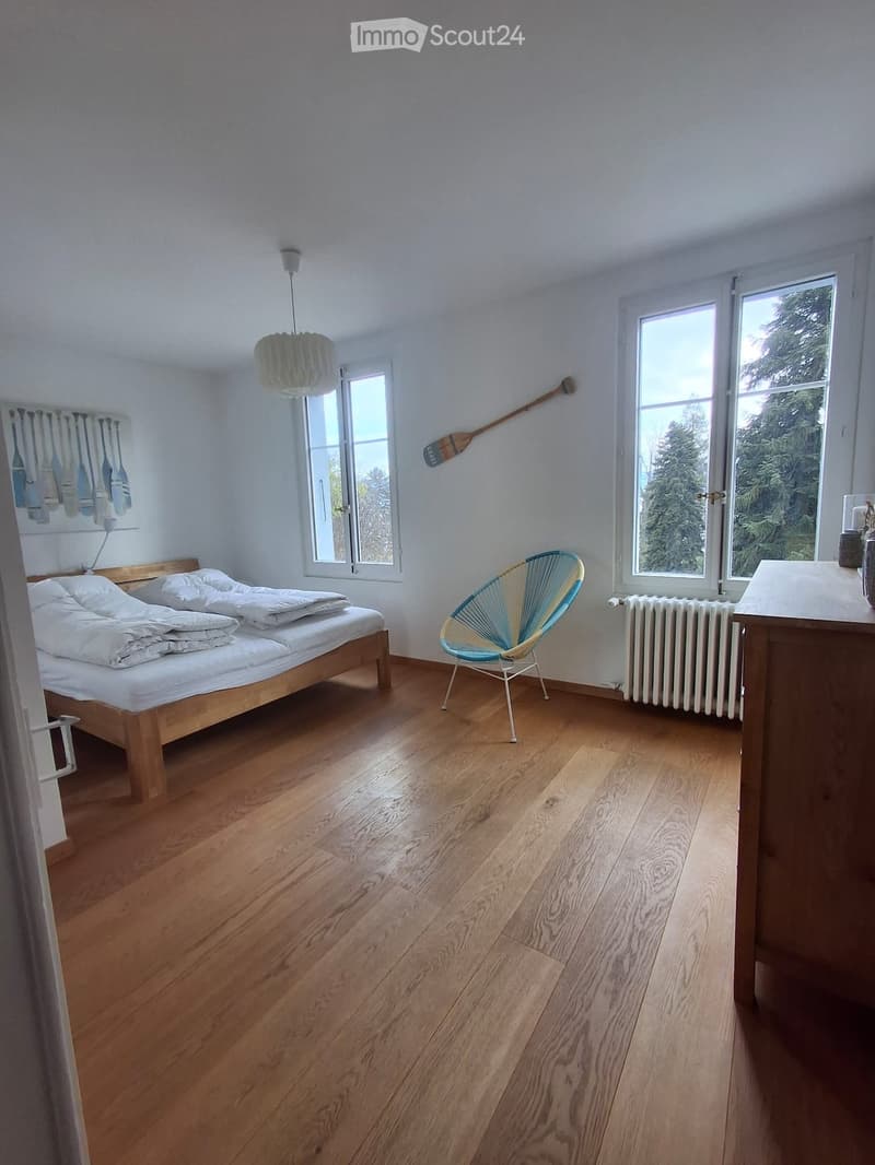 Bright bedroom with wooden flooring, large windows, a bed with white bedding, a modern chair, and a decorative oar on the wall.