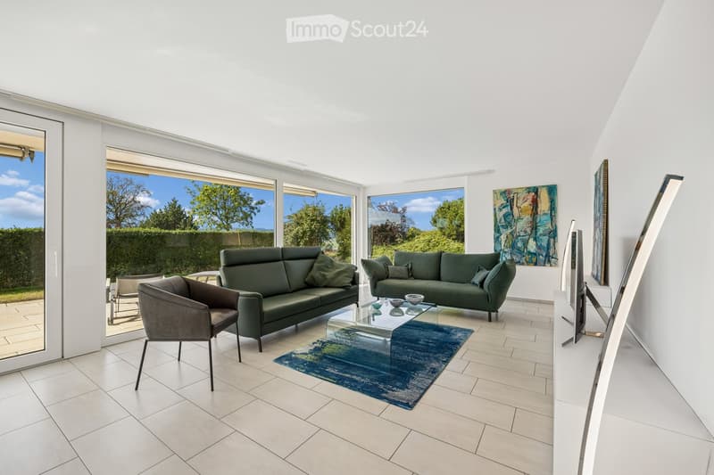 Bright living room with white walls, tiled floor, large windows offering a view of greenery, two green sofas, an armchair, a glass coffee table, a blue rug, and a large screen.