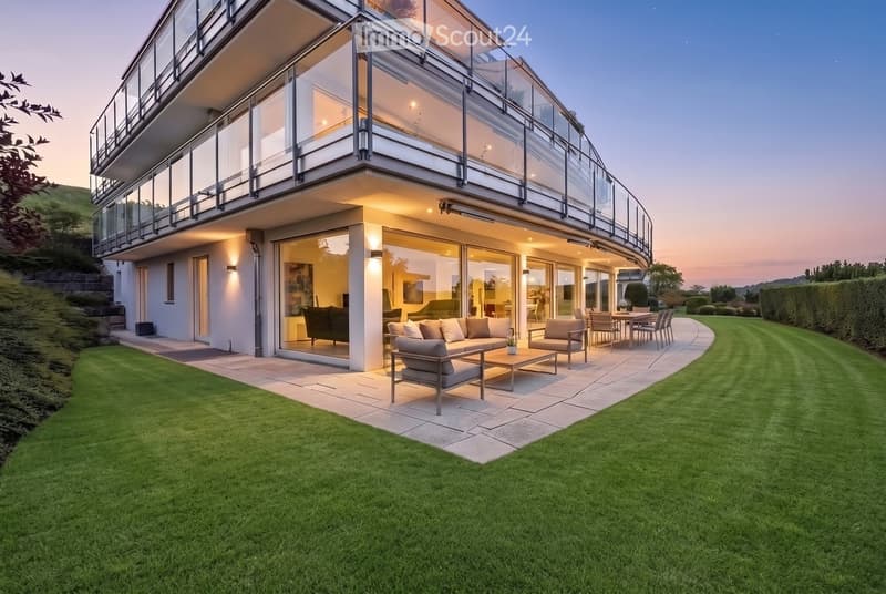 Modern house with large glass windows and a spacious outdoor seating area featuring a sofa, coffee table, and dining set, surrounded by a well-maintained lawn.
