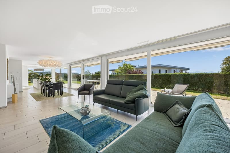 Bright living room with large windows and a view, featuring a grey sofa, coffee table, dining area, and modern decor.