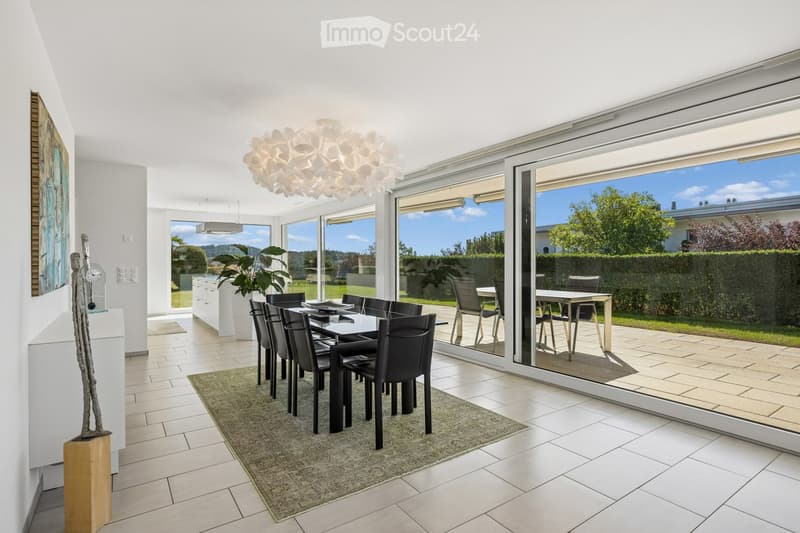 Bright dining area with modern decor, large windows offering a scenic view, a dining table with chairs, a decorative rug, a tall sculpture, and a potted plant near the kitchen.