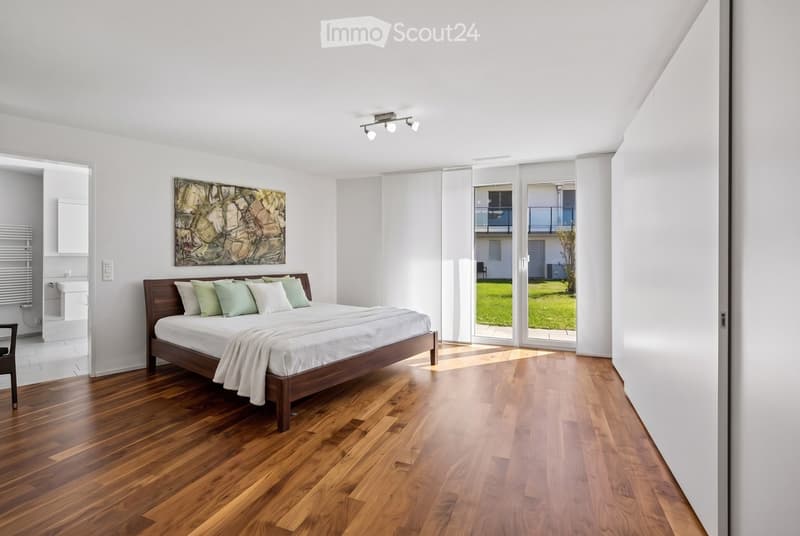 Modern bedroom with wooden flooring, a large bed with pillows and a blanket, a chair, a sink, a radiator, and a picture frame on the wall. Large sliding glass doors lead to a balcony with a view of a green lawn.