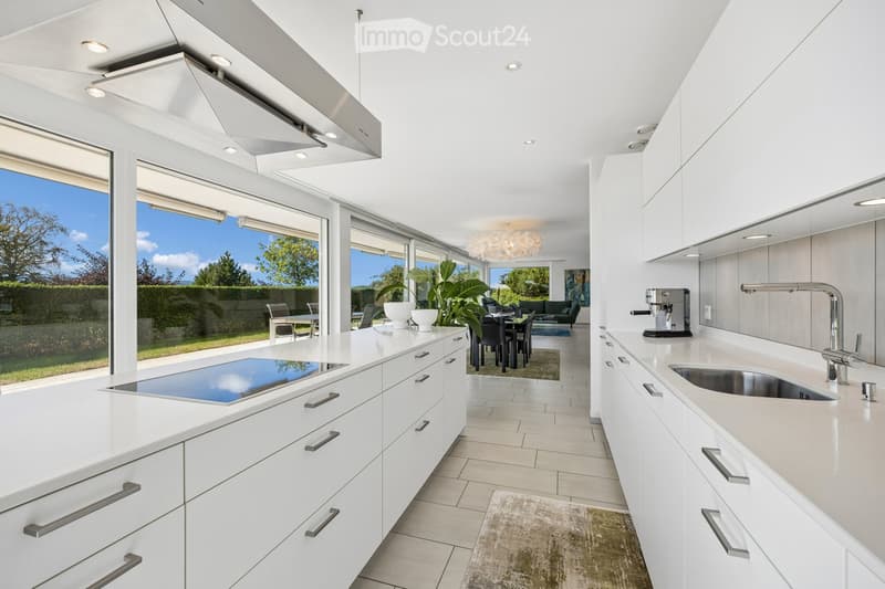Modern, open-plan kitchen with white cabinets, an induction cooktop, a stainless steel sink, and a coffee machine. Large windows offer a view of the garden, and the space is connected to a dining area with a table and chairs, and a living area with a sofa