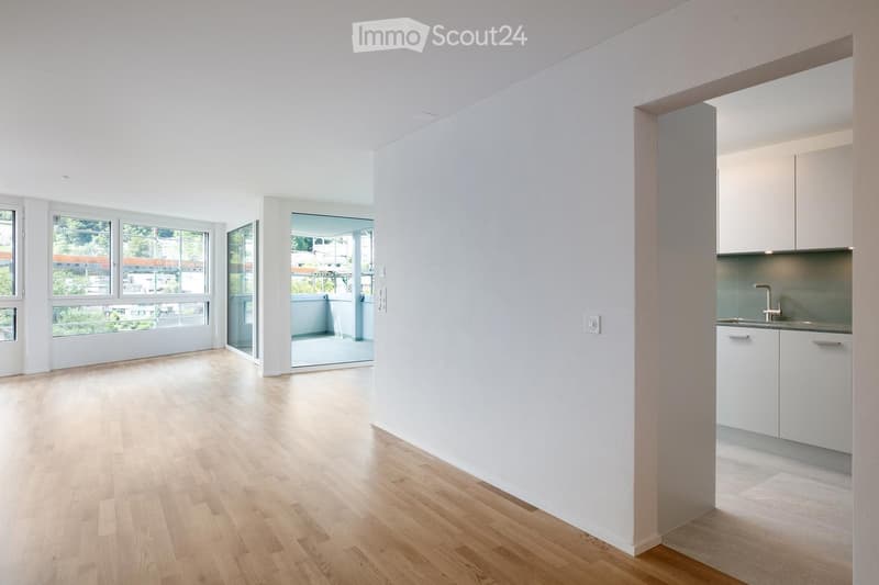 Bright, unfurnished room with light wooden flooring, white walls, large windows, and a small kitchen area with white cabinets and a sink.