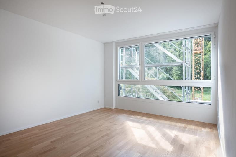 Empty room with light wooden flooring and white walls, featuring large windows with a view of an outdoor construction area.