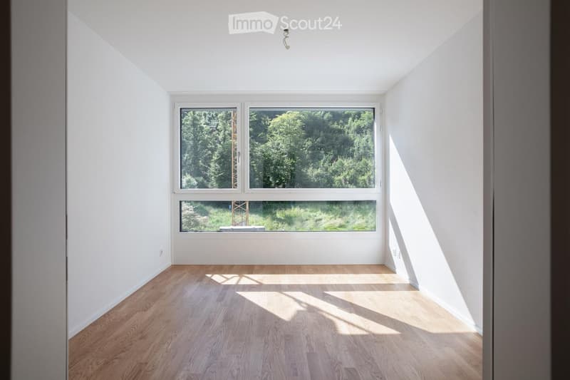 Completely empty room with white walls, wooden floor, and large windows offering a view of greenery outside.