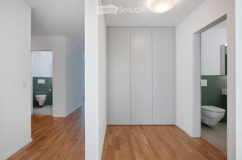 A modern, minimalist hallway with light wooden flooring, white walls, and built-in white closet doors. Two bathrooms are visible at the ends of the hallway, each with a toilet and green tiled walls.