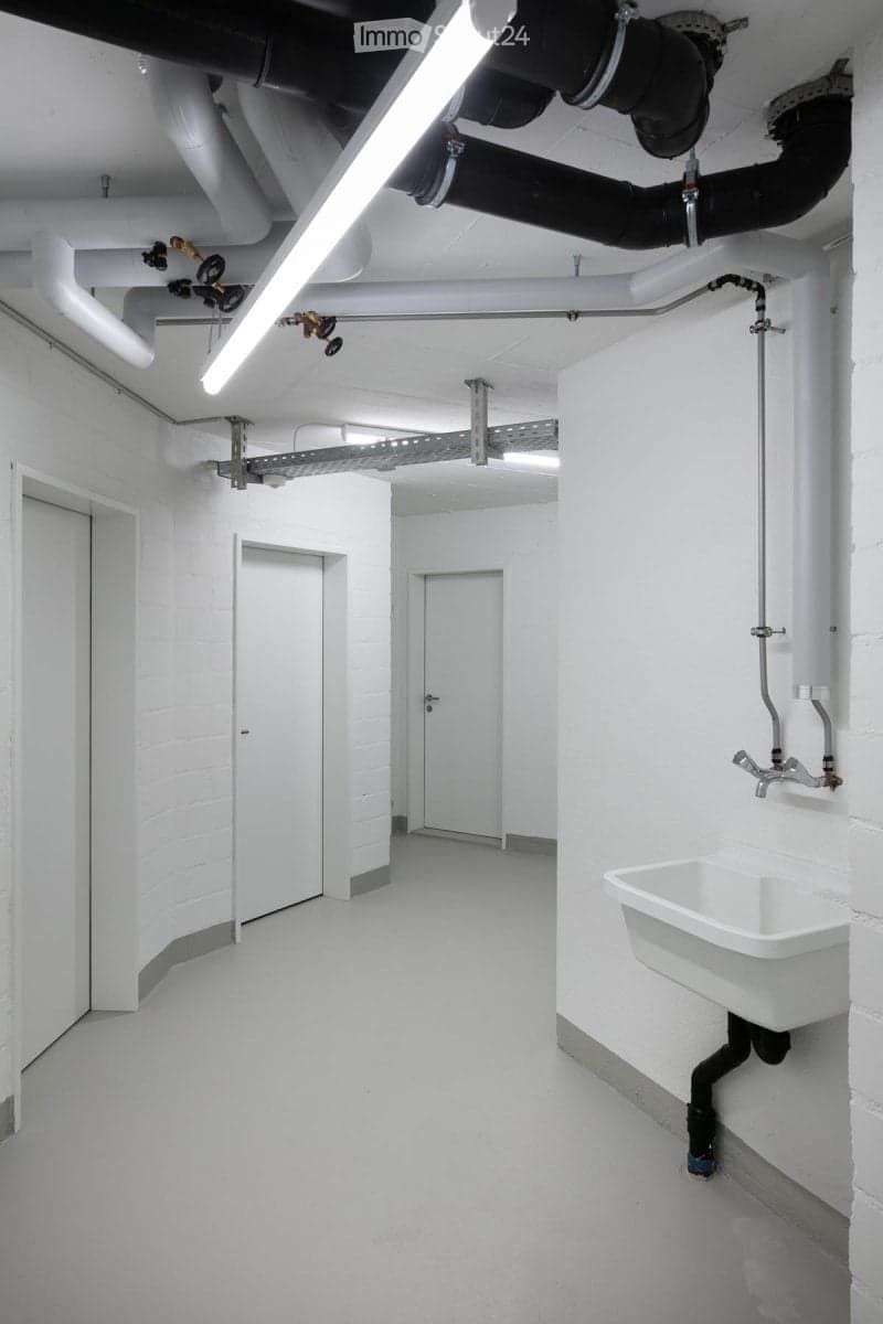 Completely empty room with bare white walls, white floor, and exposed pipes and lighting fixtures. No furniture or equipment.