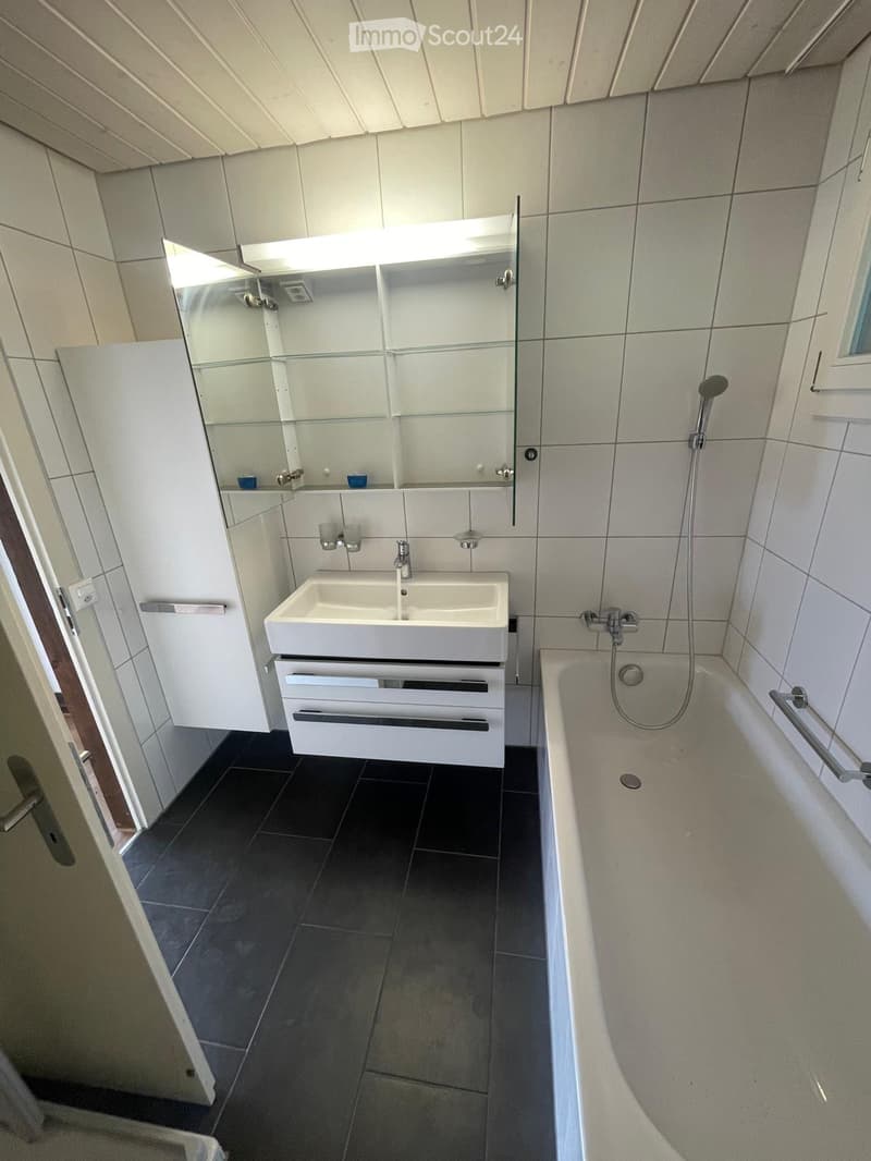 Modern bathroom with a bathtub, sink, and mirrored cabinet. Features dark tiled flooring and white tiled walls.