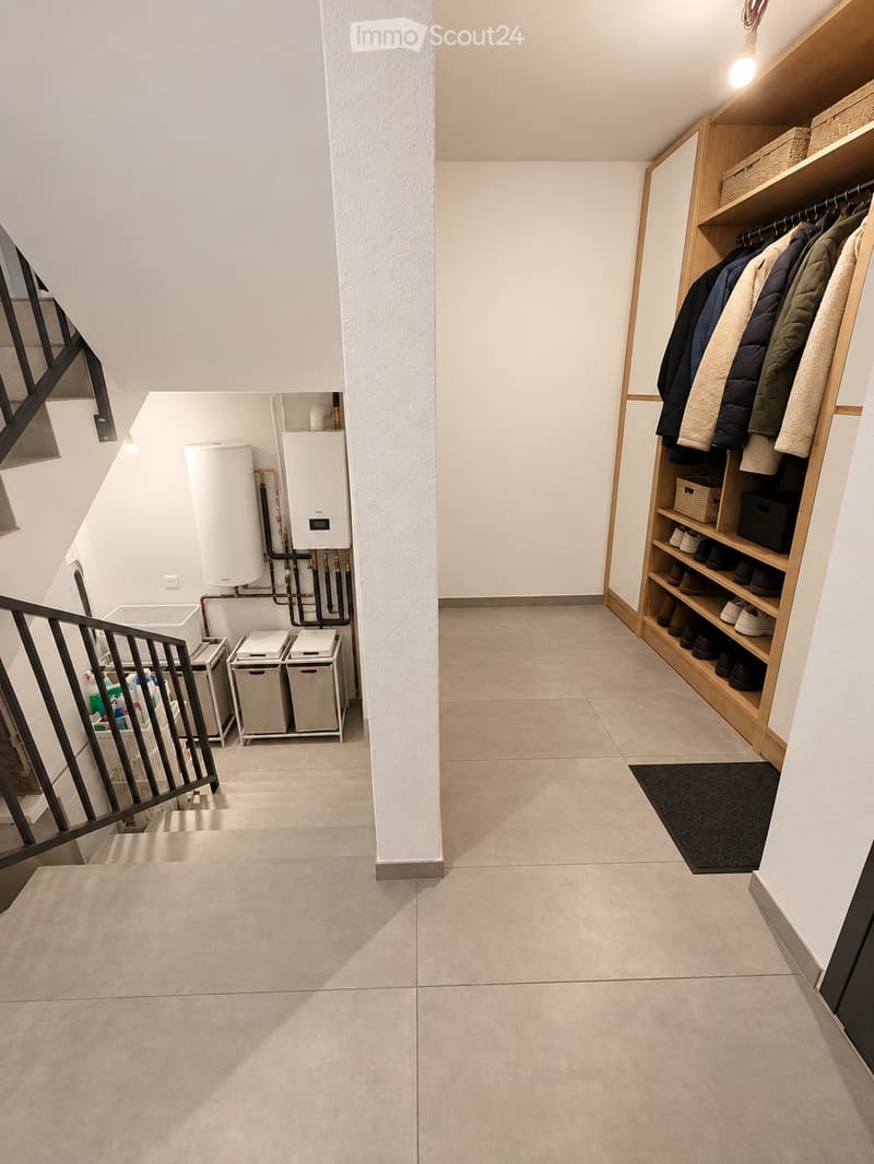 Clean, modern hallway with tiled flooring, white walls, built-in wooden coat rack, and storage units. Stairs lead upwards, with utility area visible on left side.