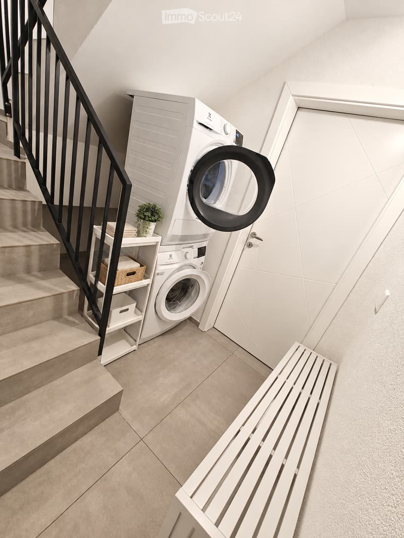 Modern bathroom with a washing machine and dryer unit, a small bench, a potted plant, and a shelf with folded towels and baskets.
