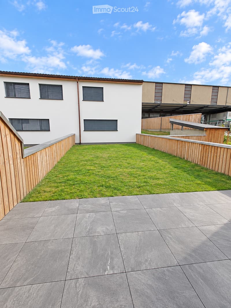 A modern outdoor patio area with a tiled floor, a small grassy lawn, wooden fencing, and a view of a contemporary building with large windows and a covered area.