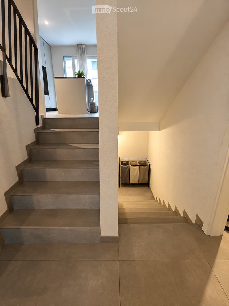 Modern hallway with tiled floors, grey carpeted stairs, black metal railing, and a small plant visible in the background.