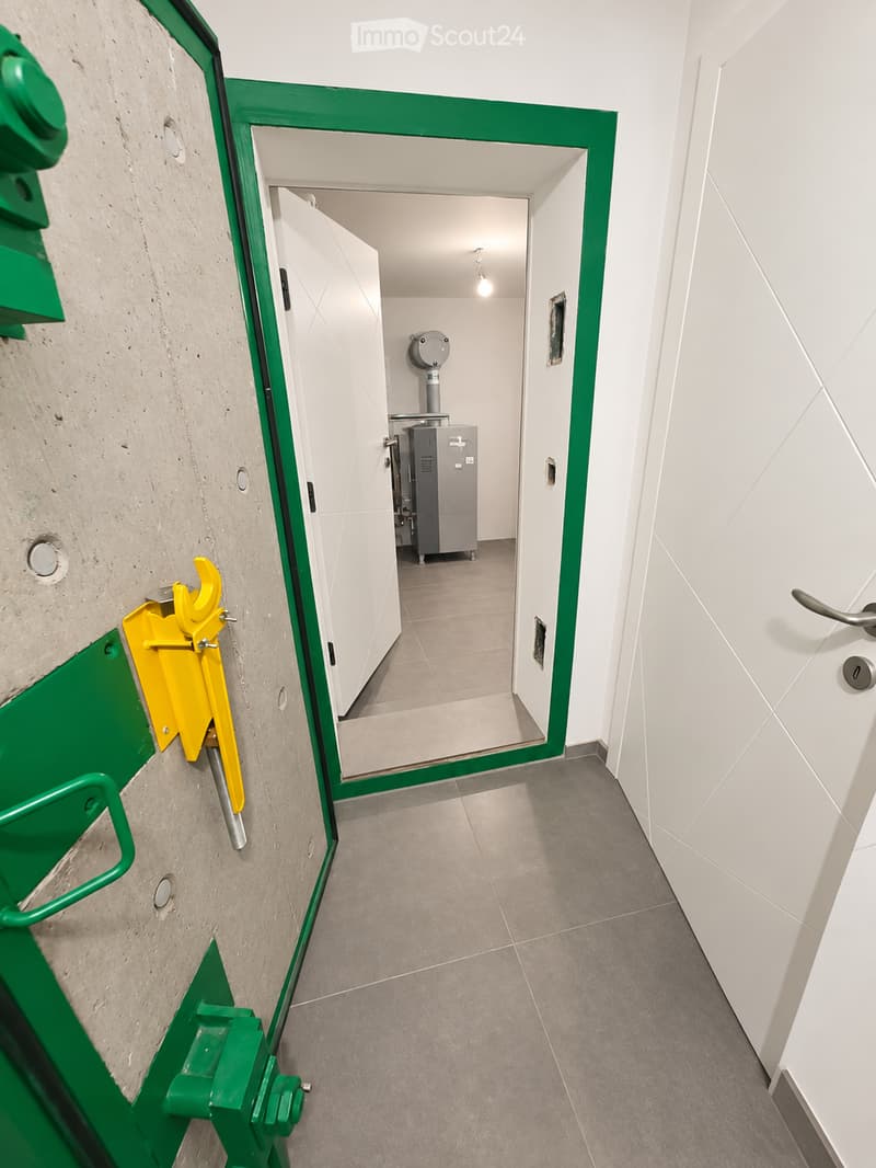 A narrow hallway with grey tiled flooring, white walls, and a green-framed doorway leading to a utility area with a grey machine and a light fixture.