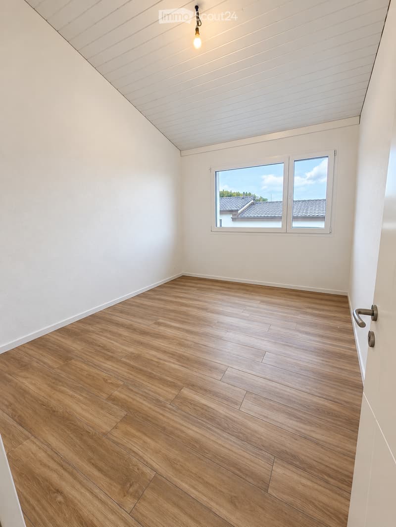 Empty room with wooden flooring, white walls, and large windows offering a view of a roof and sky.